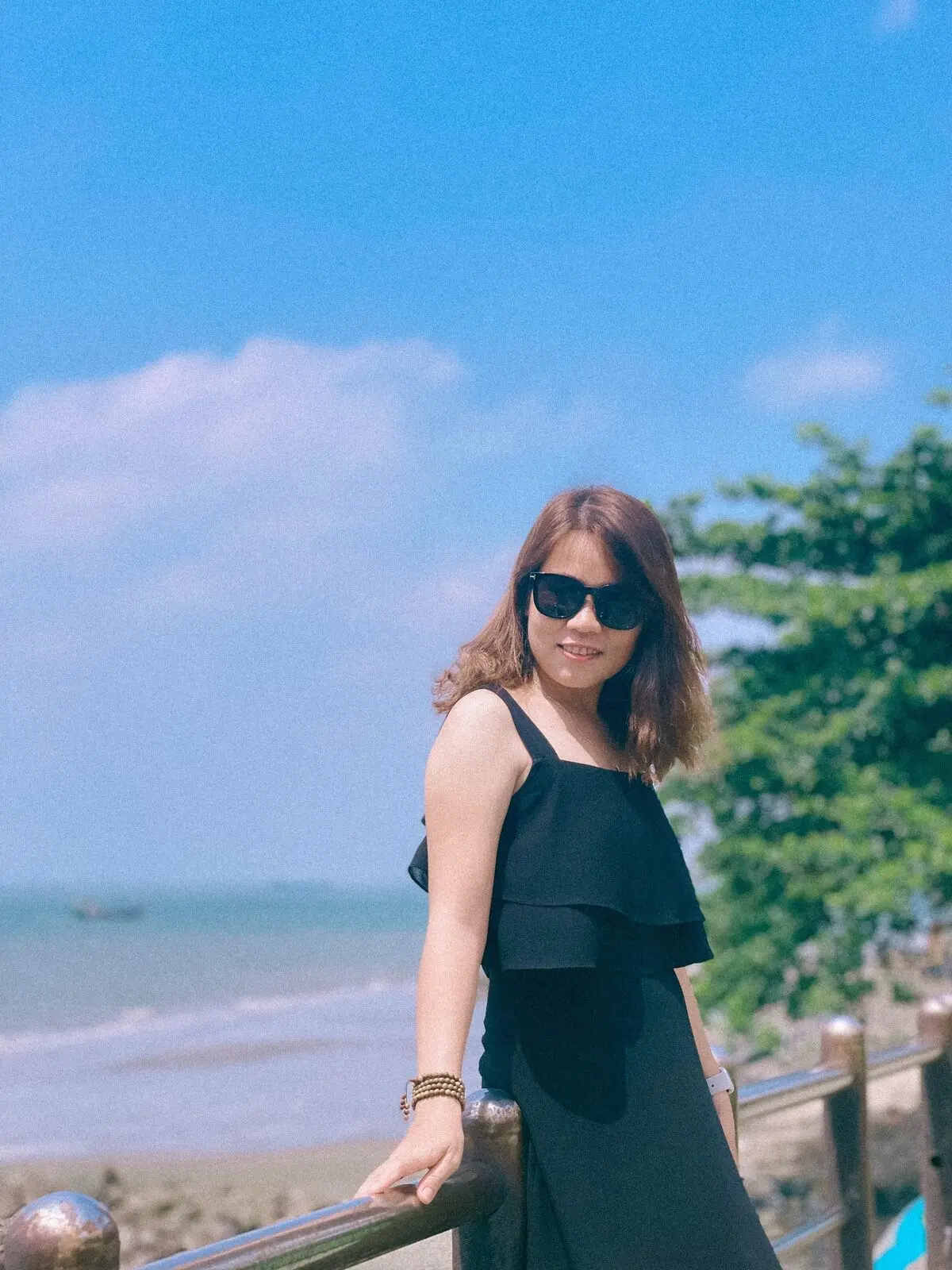 Loo đeo kính râm, mặc váy đen bên lan can nhìn ra biển