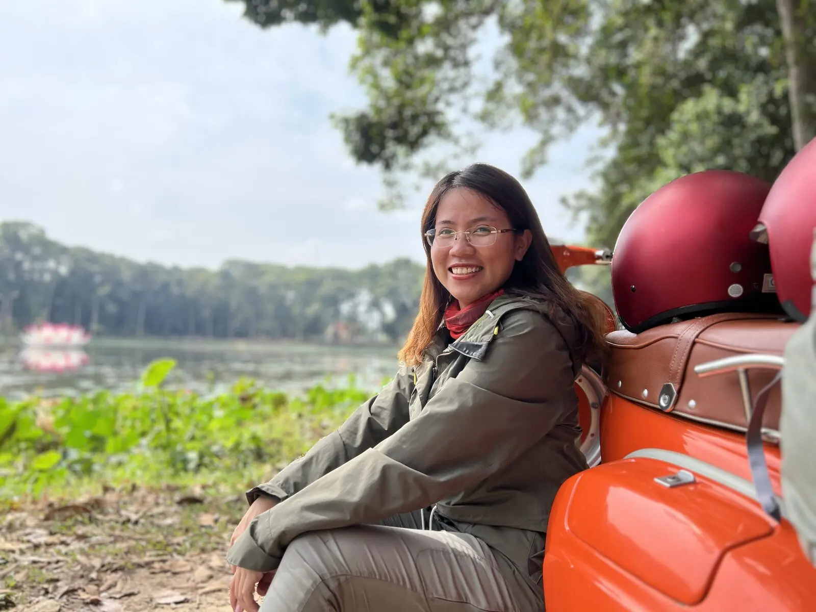 Loo ngồi tựa chiếc Vespa cam bên Ao Bà Om, mỉm cười