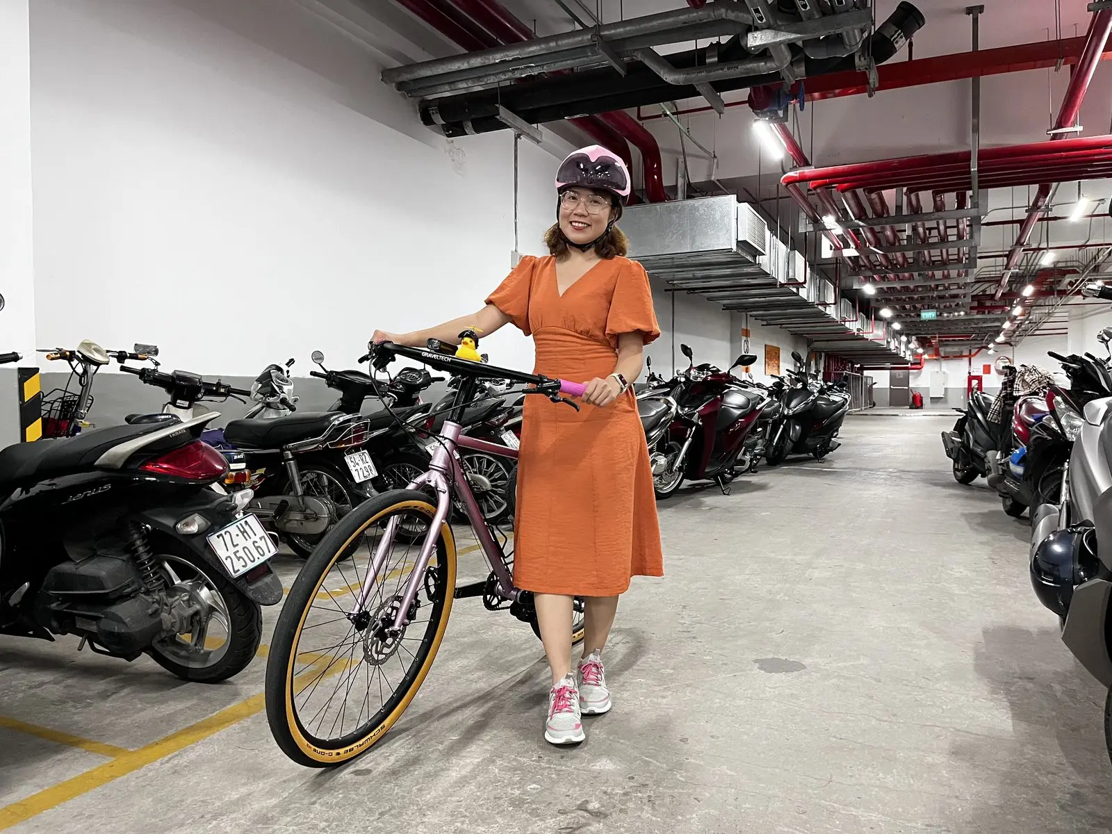Loo mặc váy cam đứng cạnh chiếc xe đạp trong hầm xe