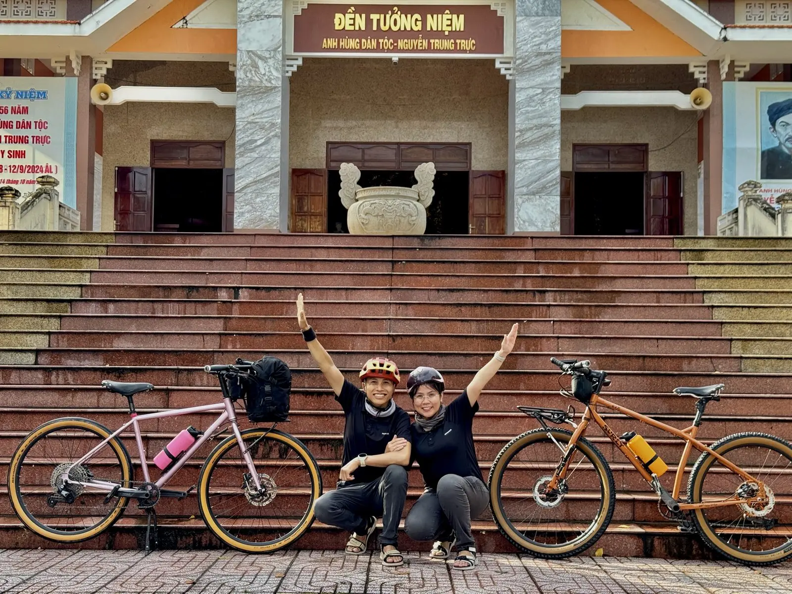 Loo và Boo giơ tay trên bậc thềm đền tưởng niệm Nguyễn Trung Trực, hai chiếc xe đạp hai bên