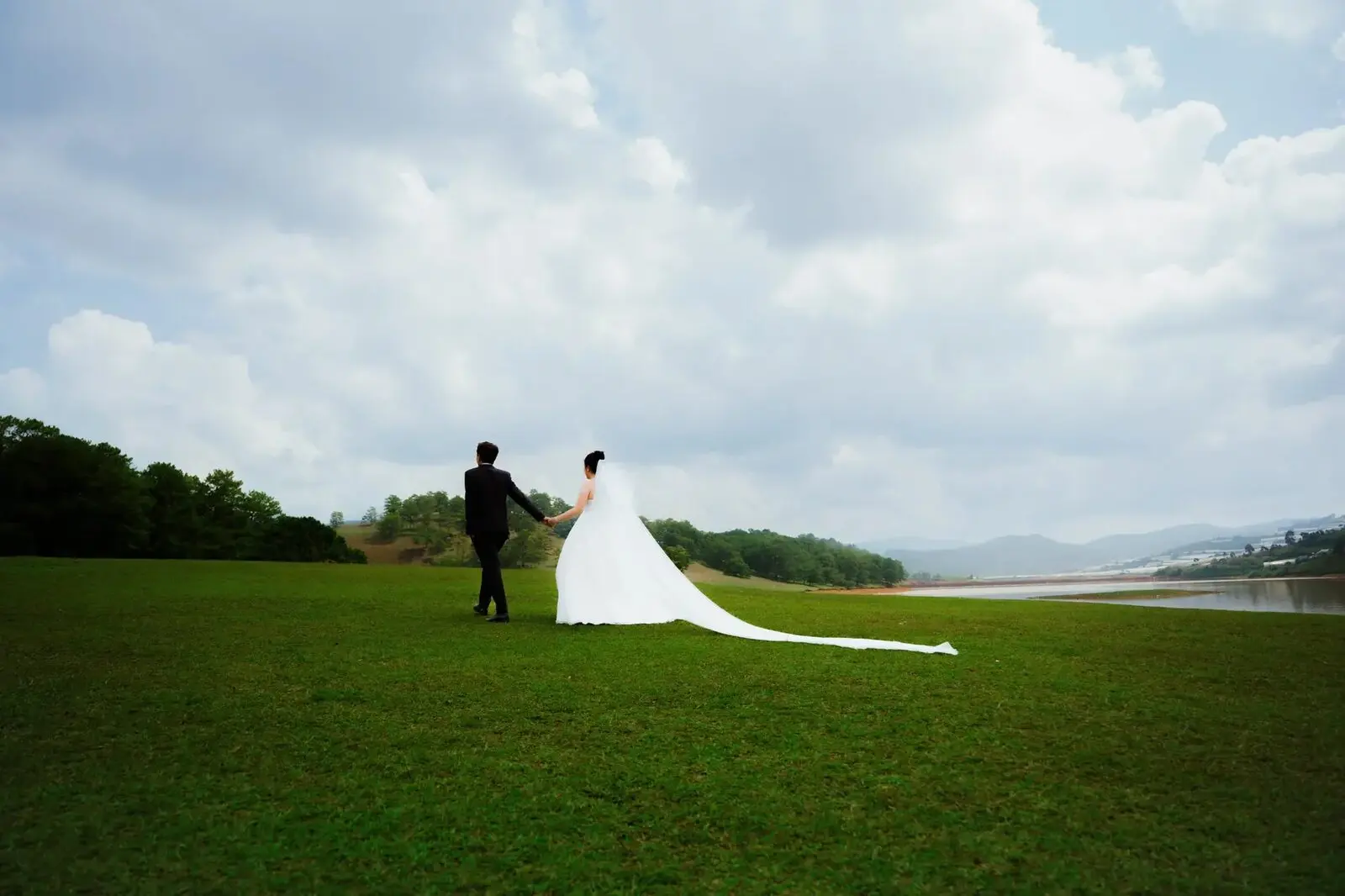 Loo và Boo nắm tay đi trên đồi cỏ xanh, váy cưới dài phía sau